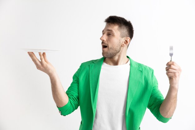 Chico caucásico atractivo sonriente joven con plato vacío y tenedor aislado sobre fondo gris. Copie el espacio y simule. Fondo de plantilla en blanco.