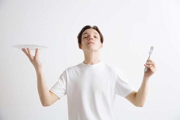 Chico caucásico atractivo sonriente joven con plato vacío y tenedor aislado sobre fondo gris. Copie el espacio y simule. Fondo de plantilla en blanco.