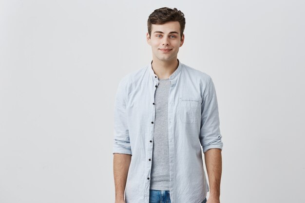 Chico caucásico contento alegre con ojos azules y cabello oscuro vestido con elegante camisa azul sonriendo de buen humor mientras posando en estudio. Concepto de expresiones faciales