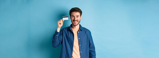 Chico caucásico feliz sonriendo mostrando tarjeta de crédito de plástico de pie en ropa casual sobre fondo azul