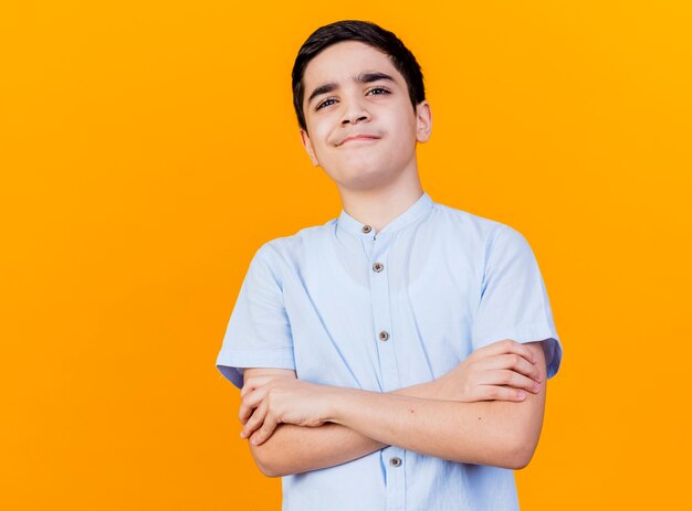 Chico caucásico joven pensativo de pie con postura cerrada mirando a cámara aislada sobre fondo naranja con espacio de copia