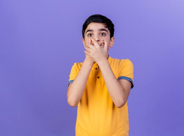 Chico caucásico joven sorprendido mirando recto manteniendo las manos en la boca aisladas sobre fondo púrpura con espacio de copia