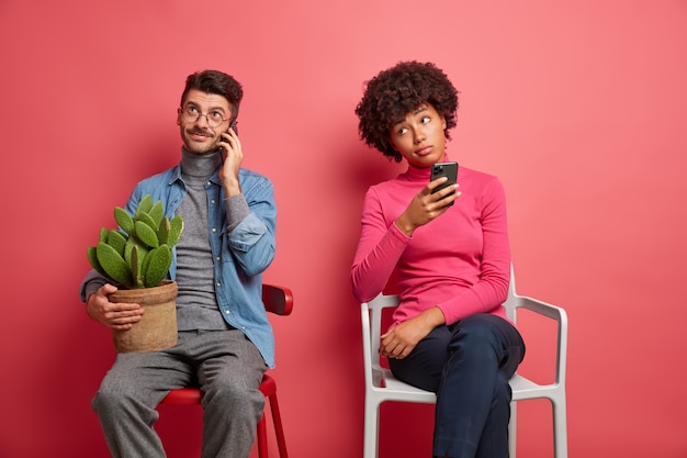 Chico caucásico tiene conversación telefónica sostiene una olla de cactus y posa en casa en una silla. Aburrida mujer de piel oscura tiene celular y piensa qué respuesta dar. Personas y tecnologías modernas