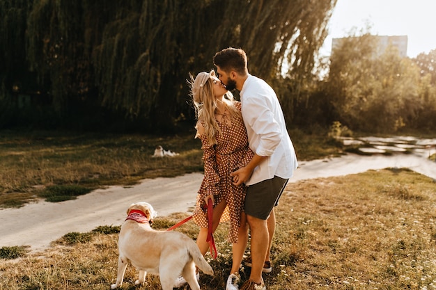 Chico y chica se besan contra el fondo de sauce. Pareja romántica tiene paseo matutino con su amado perro.