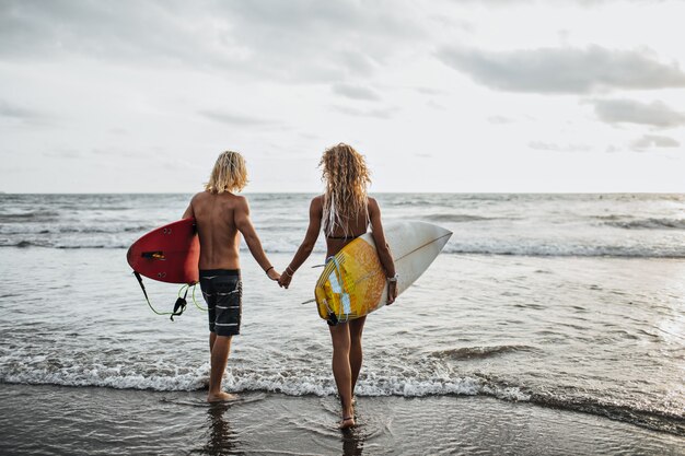 Chico y chica con cabello ondulado van de la mano al mar y sostienen tablas de surf