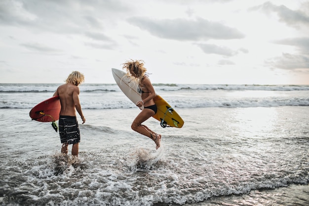 Chico y chica esbeltos saltan en agua de mar y sostienen tablas de surf