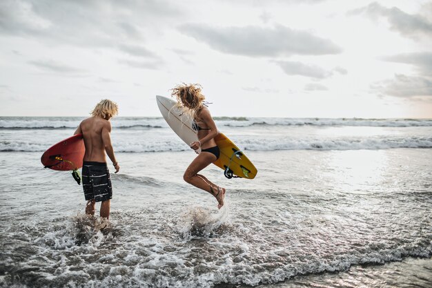 Chico y chica esbeltos saltan en agua de mar y sostienen tablas de surf