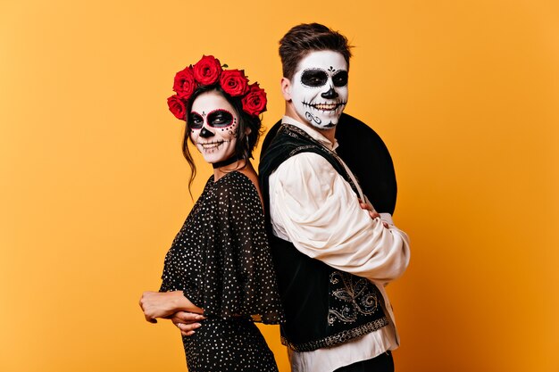 Chico y chica positivos sonríen sinceramente. Foto de pareja con maquillaje de Halloween de buen humor en pared naranja.
