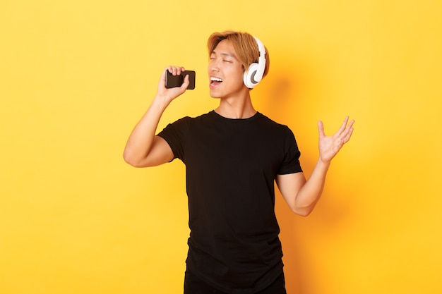 Chico coreano atractivo alegre en auriculares, jugando aplicación de karaoke, cantando en el micrófono del teléfono móvil, pared amarilla de pie