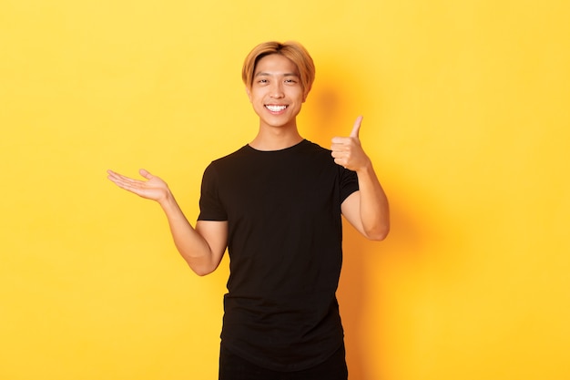 Chico coreano atractivo satisfecho y feliz sonriendo, mostrando el pulgar hacia arriba en aprobación con regocijo, sosteniendo algo en la mano, de pie en la pared amarilla
