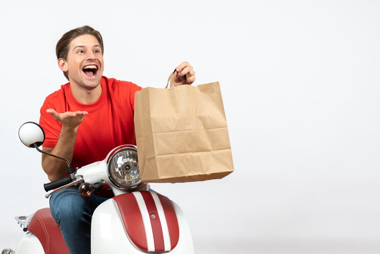 Foto gratuito chico curioso mensajero sonriente joven en uniforme rojo sentado en scooter sosteniendo una bolsa de papel en la pared blanca