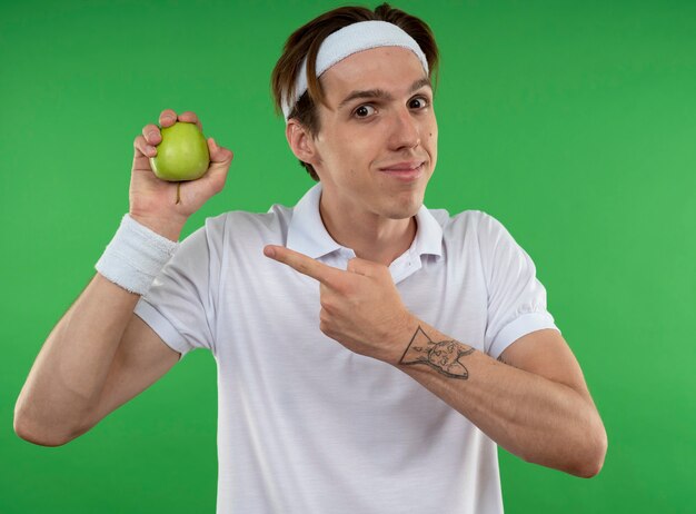 Chico deportivo joven complacido con diadema con muñequera sosteniendo y apunta a apple aislado en la pared verde