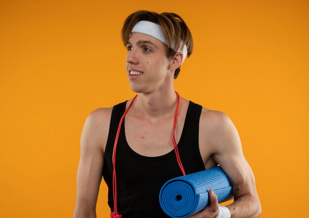 Foto gratuita chico deportivo joven complacido mirando de lado con saltar la cuerda en el cuello sosteniendo la estera de yoga aislada en la pared naranja