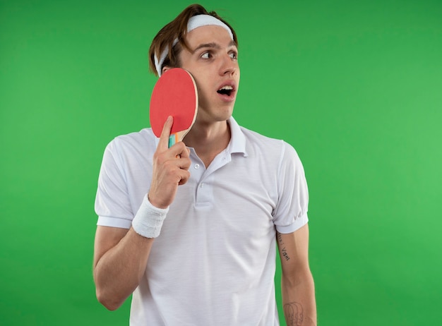Foto gratuita chico deportivo joven sorprendido mirando de lado con diadema con muñequera poniendo raqueta de ping pong en la cara aislada en la pared verde