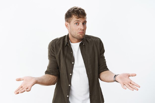 Chico despistado encogiéndose de hombros como inconsciente. Retrato de hombre rubio lindo confundido tomados de la mano de lado aturdido, frunciendo los labios y levantando las cejas cuestionado contra la pared gris