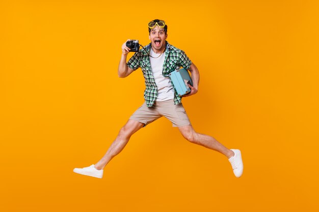 Chico divertido turista en traje de verano con cámara retro y maleta azul. Hombre con máscara de buceo saltando sobre el espacio naranja.