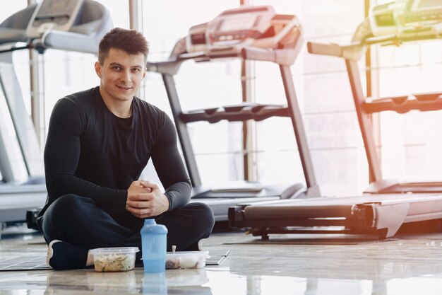 Chico elegante en el gimnasio, relajarse en el piso y comer alimentos saludables.