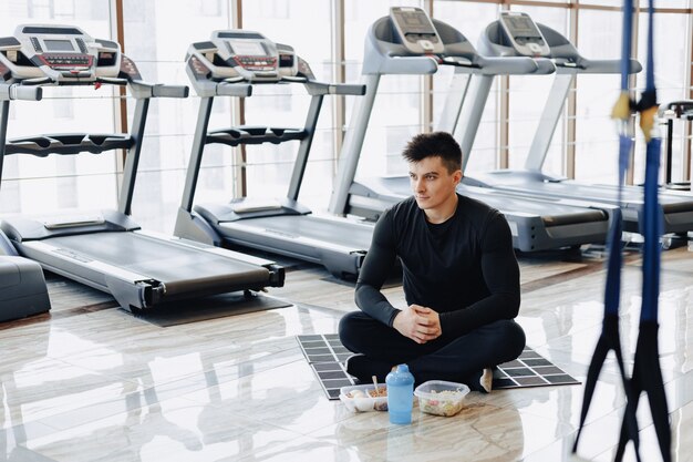 Chico elegante en el gimnasio, relajarse en el piso y comer alimentos saludables.