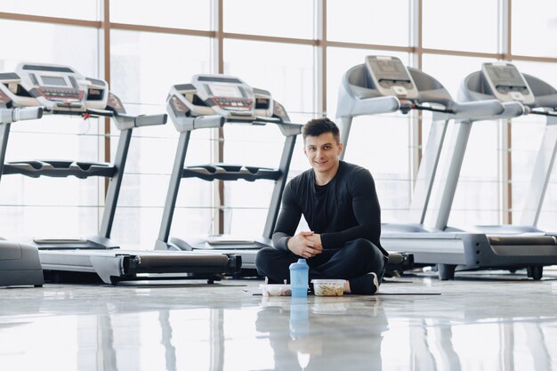 Chico elegante en el gimnasio, relajarse en el piso y comer alimentos saludables.