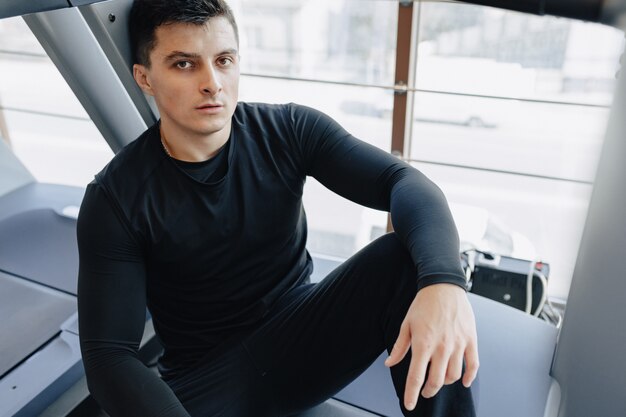 Chico elegante en el gimnasio se sienta descansando en la cinta. Estilo de vida saludable.