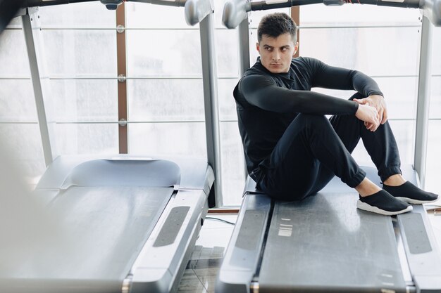 Chico elegante en el gimnasio se sienta descansando en la cinta. Estilo de vida saludable.