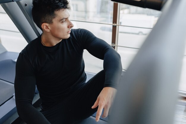 Chico elegante en el gimnasio se sienta descansando en la cinta. Estilo de vida saludable.