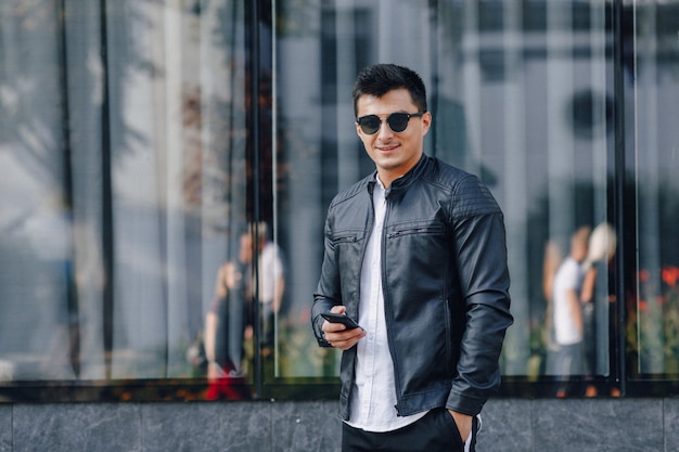 Chico elegante joven con gafas en chaqueta de cuero negro con teléfono sobre fondo de cristal