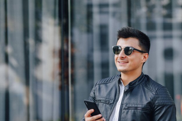 Chico elegante joven con gafas en chaqueta de cuero negro con teléfono sobre fondo de cristal