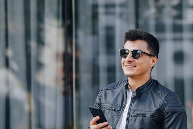 Chico elegante joven con gafas en chaqueta de cuero negro con teléfono sobre fondo de cristal