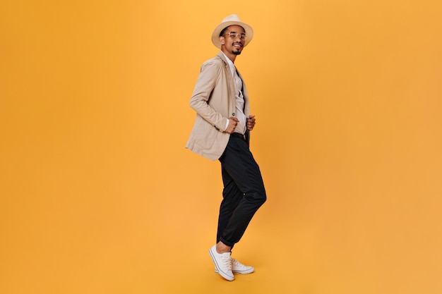 Chico elegante en pantalones negros y chaqueta beige posando sobre fondo naranja Joven en traje y sombrero mirando a la cámara en aislado