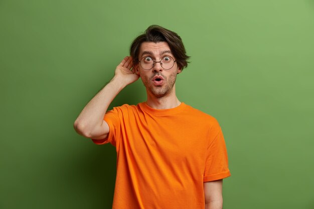 Un chico emocional aturdido escucha a escondidas información, mantiene la mano cerca del oído, se sorprende al escuchar algo inesperado, se sorprende por los chismes, usa gafas y una camiseta naranja, se para contra la pared verde