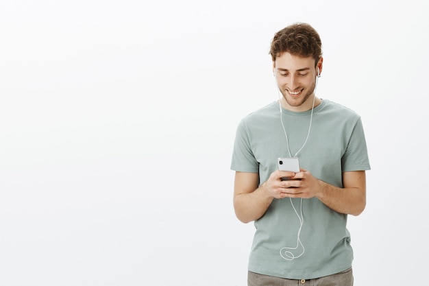 Chico escuchando música en auriculares. Retrato de hombre en camiseta, sosteniendo smartphone y sonriendo a la pantalla
