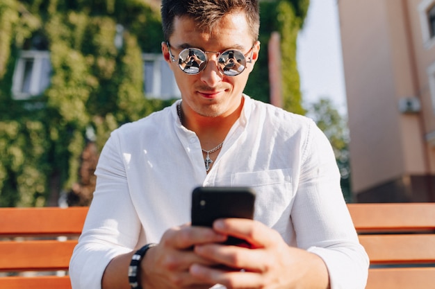 Foto gratuita chico con estilo joven en camisa con teléfono en banco en día cálido y soleado al aire libre