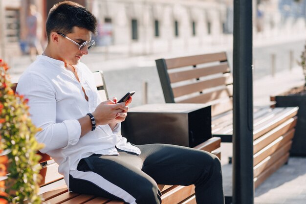 Chico con estilo joven en camisa con teléfono en el banco en un día cálido y soleado al aire libre