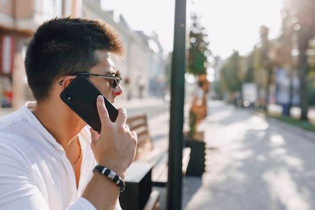 Chico con estilo joven en camisa con teléfono en banco en día soleado al aire libre