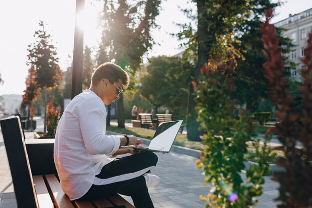 Chico con estilo joven en camisa con teléfono y portátil en banco en día soleado al aire libre