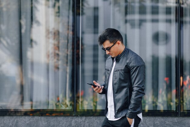Chico con estilo joven con gafas en chaqueta de cuero negro con teléfono en superficie de cristal