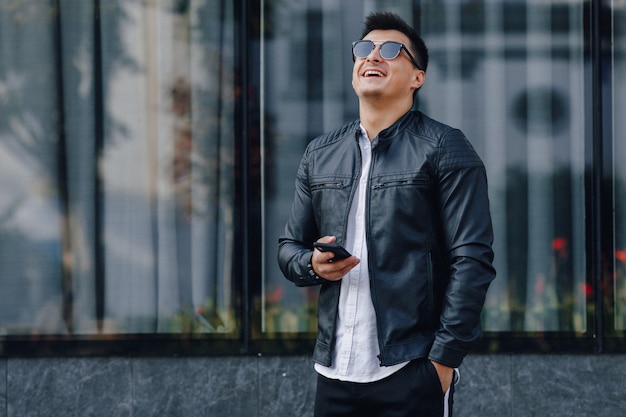 Chico con estilo joven con gafas en chaqueta de cuero negro con teléfono en superficie de cristal