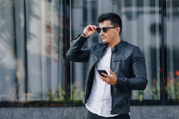 Chico con estilo joven con gafas en chaqueta de cuero negro con teléfono en superficie de cristal