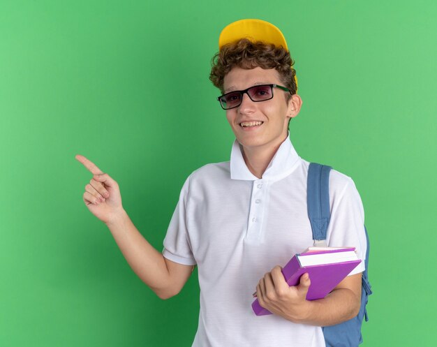 Chico estudiante con camisa blanca y gorra amarilla con gafas con mochila sosteniendo cuadernos mirando a la cámara sonriendo alegremente apuntando con el dedo índice hacia un lado