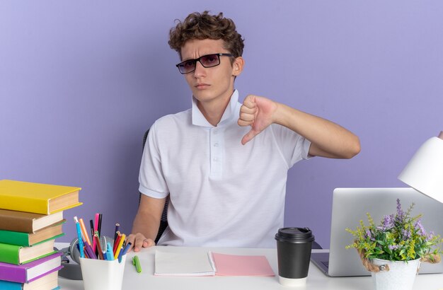 Chico estudiante en camisa polo blanca con gafas sentado en la mesa con libros mirando a la cámara con cara seria apuntando a sí mismo sobre fondo azul.