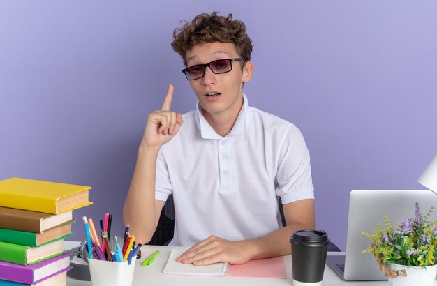 Chico estudiante en camisa polo blanca con gafas sentado en la mesa con libros mirando sorprendido y feliz de tener una nueva idea sobre la superficie azul