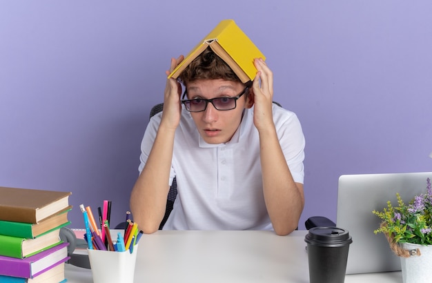 Foto gratuita chico estudiante con camisa polo blanca con gafas sentado en la mesa sosteniendo un libro sobre su cabeza mirando confundido sobre la superficie azul