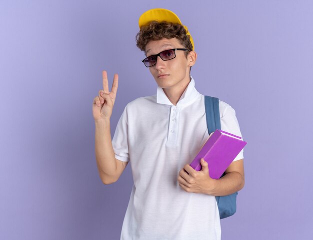 Chico estudiante con camisa polo blanca y gorra amarilla con gafas con mochila sosteniendo el cuaderno mirando a la cámara con cara seria mostrando signo v de pie sobre fondo azul