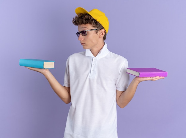 Chico estudiante en camisa polo blanca y gorra amarilla con gafas sosteniendo cuadernos