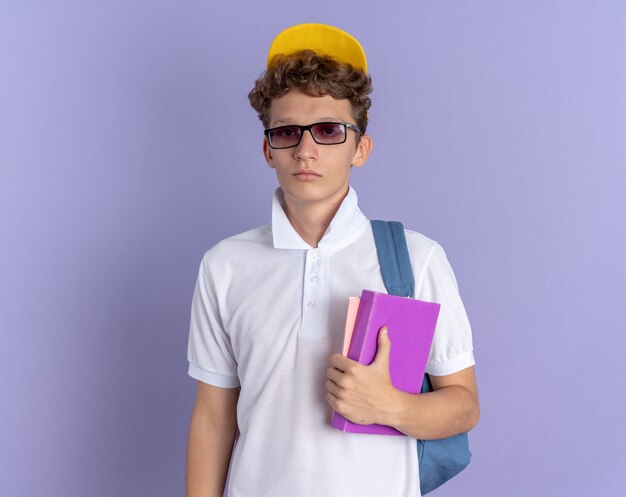 Chico estudiante en polo blanco y gorra amarilla con gafas con mochila sosteniendo cuadernos mirando a la cámara con cara seria