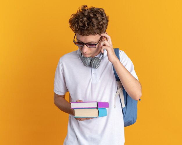 Chico estudiante en ropa casual con auriculares con gafas con mochila sosteniendo libros