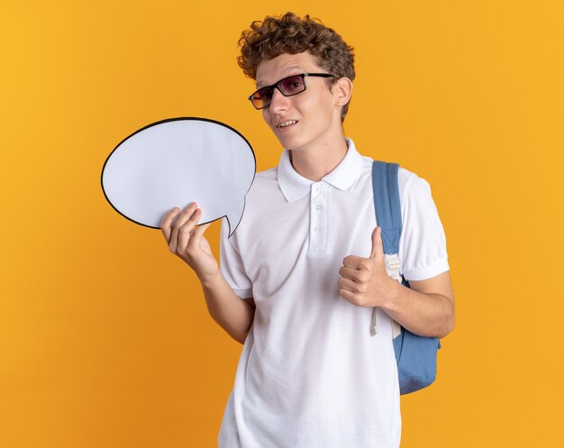 Chico estudiante en ropa casual con gafas con mochila con cartel de burbujas de discurso en blanco mirando a la cámara mostrando los pulgares para arriba sonriendo confiado de pie sobre fondo naranja