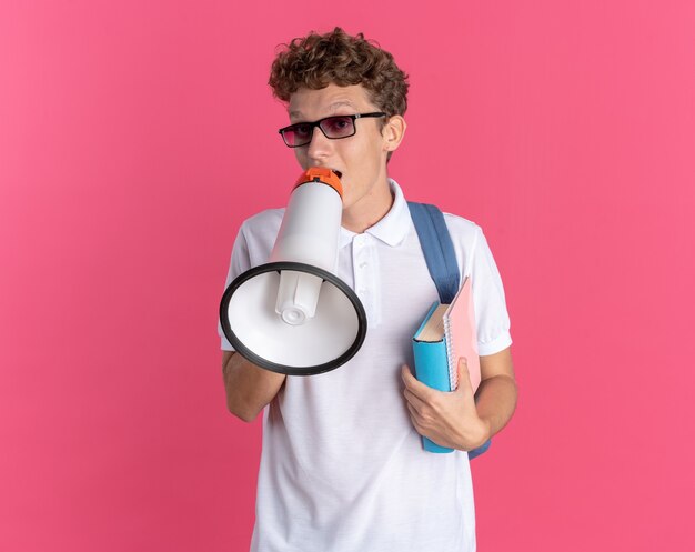 Chico estudiante en ropa casual con gafas con mochila sosteniendo cuadernos gritando al megáfono de pie sobre fondo rosa