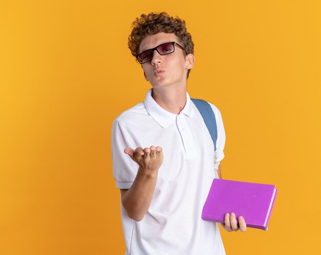 Chico estudiante en ropa casual con gafas con mochila sosteniendo el libro que sopla un beso sosteniendo la mano frente a él de pie sobre fondo naranja